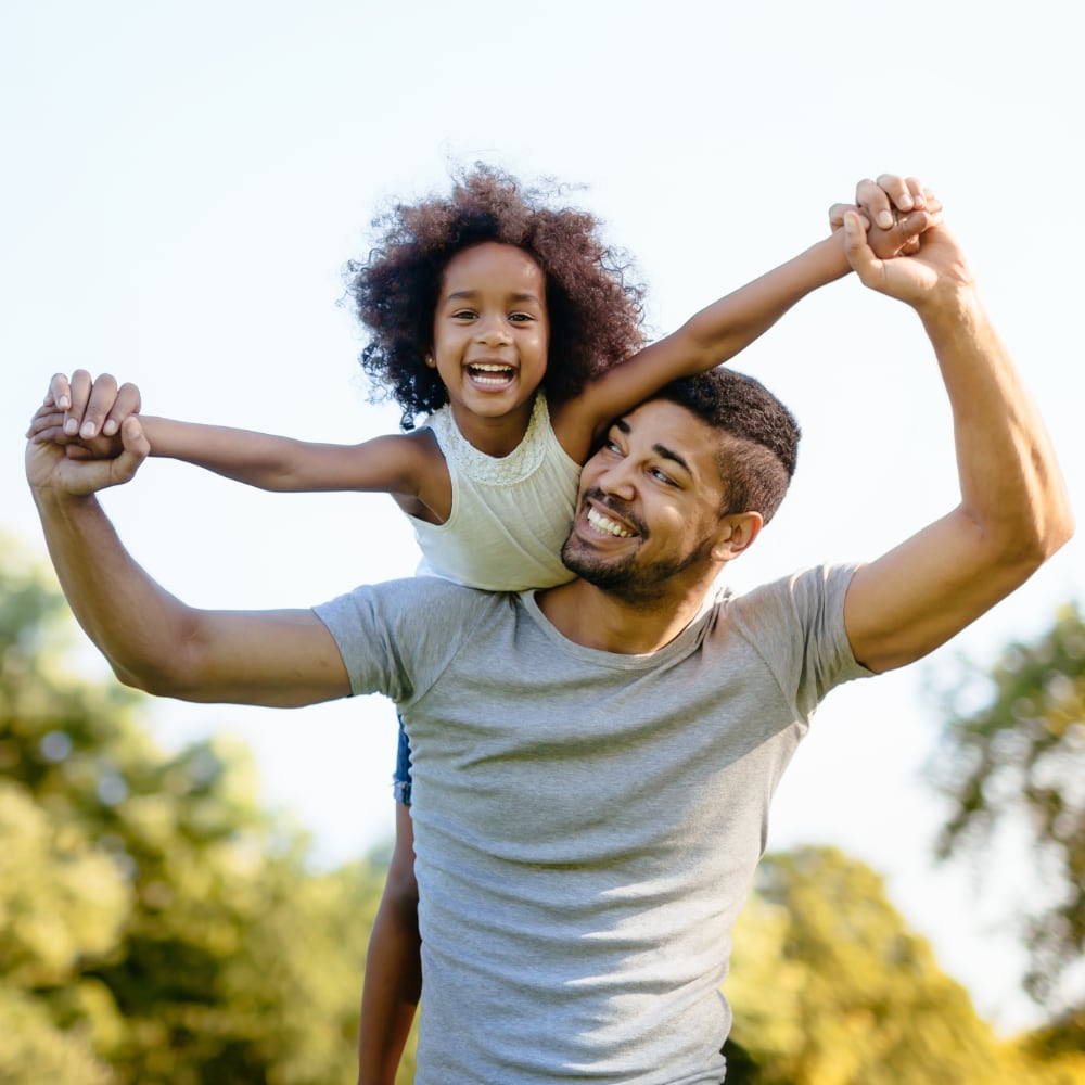 A Black father holds his smiling daughter on his shoulders at the park